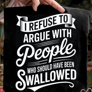 Pode incluir: Camiseta preta com o texto branco "I REFUSE TO ARGUE WITH PEOPLE WHO SHOULD HAVE BEEN SWALLOWED." O texto está em uma fonte em negrito e estilizada. A camiseta é segurada por uma mão.