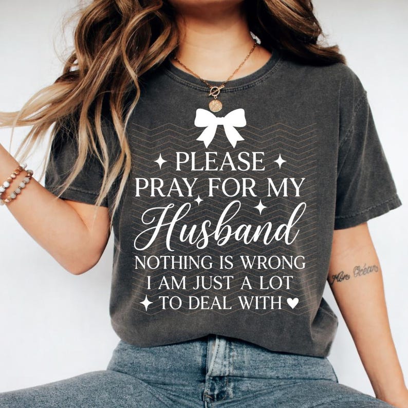 May include: Dark grey t-shirt with the text "PLEASE PRAY FOR MY Husband" in white script. The shirt also includes the phrase "NOTHING IS WRONG I AM JUST A LOT TO DEAL WITH" in white. A white bow graphic is above the text.