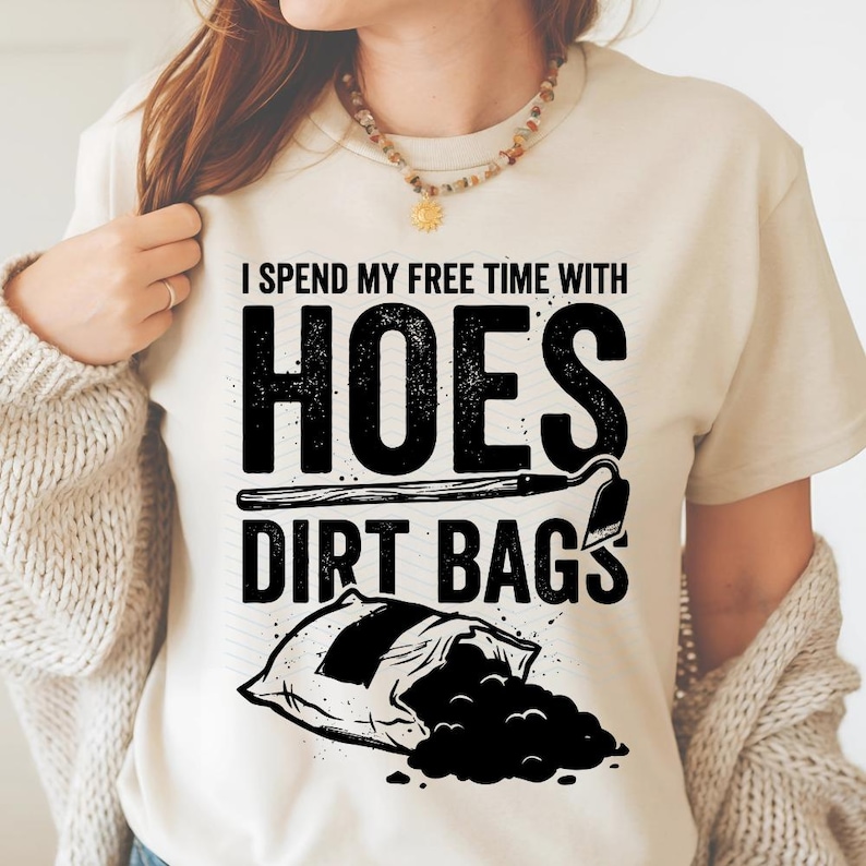 May include: A cream-coloured t-shirt featuring the text "I Spend My Free Time With Hoes Dirt Bags" in black. The design includes a garden hoe and a bag of soil, ideal for those who enjoy gardening.