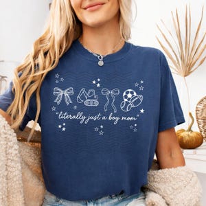 Puede incluir: Camiseta azul marino con texto e ilustraciones en blanco. El texto dice "literally just a boy mom" con imágenes de un lazo, una excavadora, otro lazo y un balón de fútbol y rugby. La camiseta tiene cuello redondo.