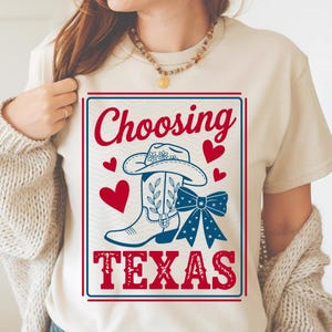 May include: Cream-colored t-shirt with a graphic design featuring a cowboy hat, a cowboy boot, and a bow, all in blue and white, with red hearts. The words "Choosing TEXAS" are printed in red.