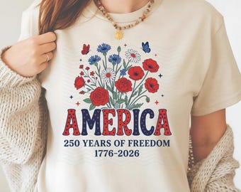 Może przedstawiać: Kremowa koszulka z kwiatowym wzorem z czerwonymi różami, niebieskimi chabrami i białymi stokrotkami. Słowo "AMERICA" jest w czerwono-niebieskie grochy, z tekstem "250 YEARS OF FREEDOM 1776-2026" poniżej.