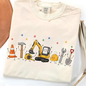 Könnte beinhalten: Ein cremefarbenes T-Shirt mit einem Grafikdruck von Cartoon-Bauwerkzeugen und -geräten, darunter ein Verkehrskegel, ein Hammer, ein Maßband, ein Bagger, ein Schutzhelm, ein Schraubenschlüssel und eine Schaufel, alle mit lächelnden Gesichtern.