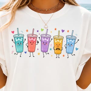 Puede incluir: Una camiseta blanca con un estampado gráfico de cinco vasos de bebidas heladas antropomórficos y coloridos en turquesa, rosa, morado, amarillo y azul. Cada vaso tiene una cara sonriente, brazos y piernas delgados, rodeado de estrellas y corazones.