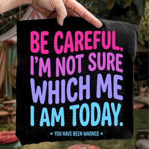 Op de afbeelding: Zwart vierkant textiel met de tekst "BE CAREFUL. I'M NOT SURE WHICH ME I AM TODAY." in roze, paars en blauw. De tekst "YOU HAVE BEEN WARNED" staat onderaan. Het textiel wordt door een hand omhoog gehouden.