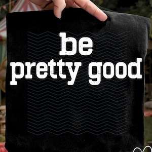 May include: Black t-shirt featuring the words "be pretty good" in white, distressed font. A dark grey chevron pattern is visible. The t-shirt is held up by a hand. The PNG logo is in the bottom right corner.