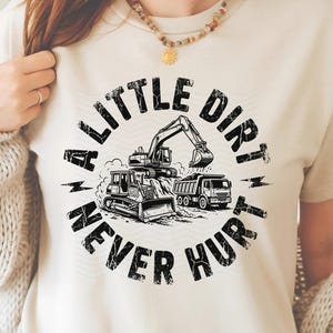 May include: Cream-colored t-shirt featuring a black and white graphic of construction vehicles, including an excavator, bulldozer, and dump truck. The text "A Little Dirt Never Hurt" is printed in a circular design around the vehicles.