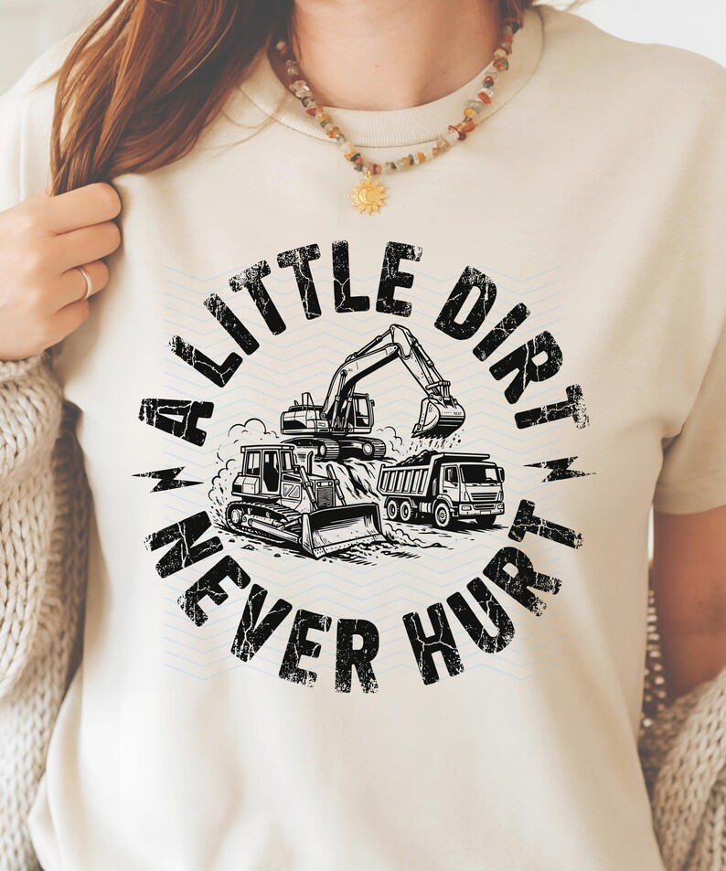 May include: Cream-colored t-shirt featuring a black and white graphic of construction vehicles, including an excavator, bulldozer, and dump truck. The text "A Little Dirt Never Hurt" is printed in a circular design around the vehicles.