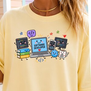 May include: A pale yellow t-shirt featuring a colorful graphic of a smiling laptop with the text SOFTWARE DEVELOPER, surrounded by animated computer monitors, a smartphone, a stack of books labeled HTML, CSS, and JavaScript, and a coffee mug.