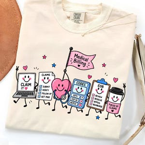 Op de afbeelding: Een crèmekleurig t-shirt met een afbeelding van stripfiguren die taken voor medische facturering voorstellen. Het ontwerp bevat een laptop, een lijst met claims, een hart met een stethoscoop, een rekenmachine met codes en een koffiebeker.