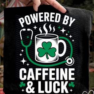 以下が含まれることがあります： 「POWERED BY CAFFEINE & LUCK」の文字が入った黒いTシャツ。デザインは、クローバーの葉、聴診器、星のアクセントが付いたコーヒーマグです。楽しい聖パトリックの日のテーマまたは医療をテーマにしたグラフィック。
