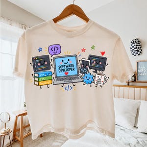 May include: A cream-colored t-shirt featuring a colorful graphic of a smiling laptop with the text "SOFTWARE DEVELOPER" and a heart. Surrounding the laptop are cartoon illustrations of a smartphone, a coffee mug labeled "CODE COFFEE REPEAT", and books labeled "HTML", "CSS", and "JavaScript".