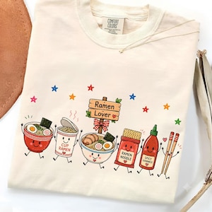 Könnte beinhalten: Ein cremefarbenes T-Shirt mit einem Grafikdruck von anthropomorphen Ramen-Motiven, darunter eine Schüssel, ein Becher, eine Nudelpackung, eine scharfe Soßenflasche und Essstäbchen, sowie ein Holzschild mit der Aufschrift Ramen Lover und bunten Sternen.