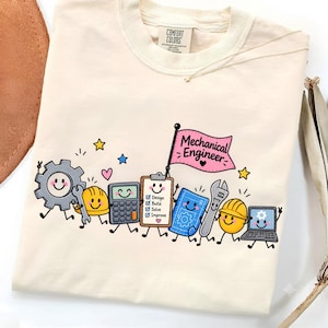 Puede incluir: Una camiseta color crema con un gráfico de herramientas de ingeniería animadas, incluyendo un engranaje, casco, calculadora, portapapeles con el texto 'Design Build Solve Improve', plano, llave inglesa, portátil y una bandera rosa que dice 'Mechanical Engineer'.