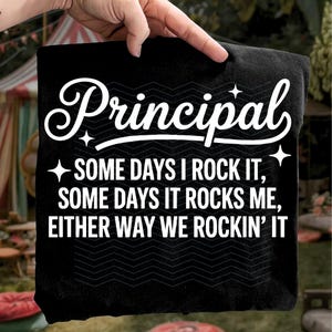 Op de afbeelding: Zwart kussen met het woord "Principal" in witte letters. Daaronder staat de tekst "SOME DAYS I ROCK IT, SOME DAYS IT ROCKS ME, EITHER WAY WE ROCKIN' IT". Het kussen wordt door een hand vastgehouden.