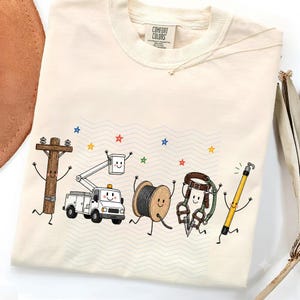 May include: A cream-coloured short-sleeve t-shirt with a Comfort Colors label at the neck. The graphic features five anthropomorphic utility items including a wooden pole, a bucket truck, a wire spool, a climbing harness, and a yellow tool.