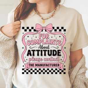 Puede incluir: Camiseta color crema con un diseño de lazo y cinta rosa. El texto dice "FOR COMPLAINTS ABOUT ATTITUDE please contact THE MANUFACTURER" en negro y rosa. El diseño incluye bordes a cuadros blancos y negros.