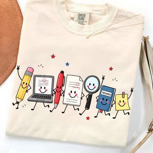 May include: A cream-colored t-shirt featuring a graphic print of anthropomorphic school supplies including a pencil, laptop, pen, paper, magnifying glass, blue book labeled THESAURUS, and a yellow sticky note, all with smiling faces.