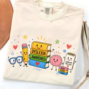 May include: A cream-coloured t-shirt featuring a colourful graphic of a stack of books labelled DYSLEXIA AWARENESS, a smiling pencil, blue glasses, a pink brain, and a notepad with the text Different Not Less and READ LEARN GROW.