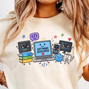 May include: A cream-colored t-shirt featuring a colorful graphic of a smiling laptop with the text SOFTWARE DEVELOPER, surrounded by a stack of books labeled HTML, CSS, and JavaScript, a smartphone, a monitor, a gear, and a coffee mug labeled CODE COFFEE REPEAT.