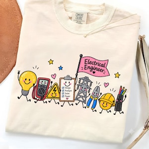 Könnte beinhalten: Ein cremefarbenes T-Shirt mit einem Grafikdruck von cartoonartigen elektrotechnischen Werkzeugen und Symbolen. Das Design umfasst eine Glühbirne, ein Multimeter, ein Warnschild, ein Klemmbrett mit Text, einen Strommast, eine Zange, einen Schutzhelm und Kabel sowie eine rosa Flagge mit der Aufschrift Electrical Engineer.