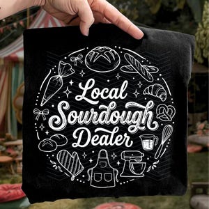 May include: Black t-shirt with a white circular design featuring baking-related illustrations. The text "Local Sourdough Dealer" is prominently displayed. The design includes images of bread, a mixer, and other baking tools.