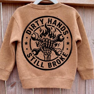 May include: A tan sweatshirt with a black graphic on the back. The graphic features a skeleton hand holding crossed wrenches, flames, and the text "DIRTY HANDS STILL BROKE" in a circular design.