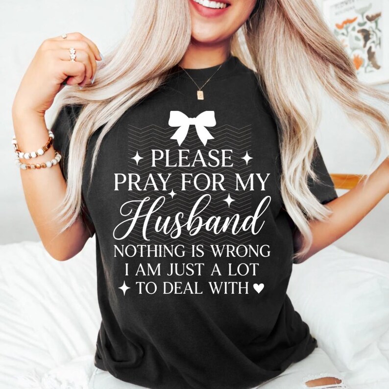 May include: Black t-shirt with the text "PLEASE PRAY FOR MY Husband NOTHING IS WRONG I AM JUST A LOT TO DEAL WITH" in white script. The shirt features a bow graphic at the top and is a casual wear item.
