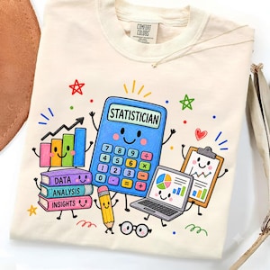 Op de afbeelding: Een crèmekleurig t-shirt met een kleurrijke afbeelding van een lachende rekenmachine met de tekst "STATISTICIAN" erboven, samen met een stapel boeken met de labels "DATA," "ANALYSIS," en "INSIGHTS," een laptop, een klembord en een potlood.