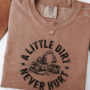 May include: A brown Comfort Colors t-shirt with the text "A Little Dirt Never Hurt" in black. The graphic features a bulldozer, excavator, and dump truck. A gold necklace with a pendant is draped across the shirt.