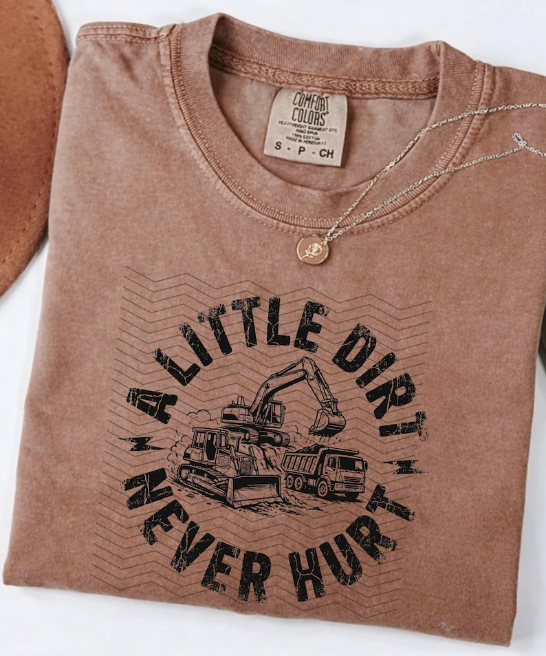 May include: A brown Comfort Colors t-shirt with the text "A Little Dirt Never Hurt" in black. The graphic features a bulldozer, excavator, and dump truck. A gold necklace with a pendant is draped across the shirt.
