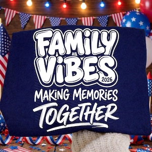 May include: A navy blue sweatshirt with the words "FAMILY VIBES 2026 MAKING MEMORIES TOGETHER" in white, outlined text. The sweatshirt is displayed with patriotic decorations in the background, including flags and balloons.