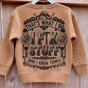 May include: A tan-colored sweatshirt with the text "THAT'S WHAT I DO, I FIX STUFF, AND I KNOW THINGS" in black. The design includes tools and gears. The sweatshirt is displayed on a white hanger.