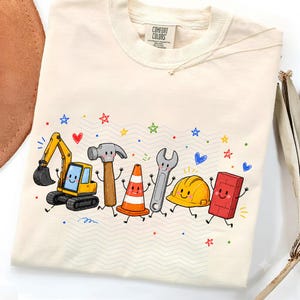Puede incluir: Una camiseta de color crema con un estampado gráfico de personajes de construcción animados, incluyendo una excavadora, un martillo, un cono de tráfico, una llave inglesa, un casco y un ladrillo. Cada elemento tiene una cara sonriente y extremidades finas.