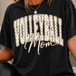 Könnte beinhalten: Ein schwarzes Kurzarm-T-Shirt mit dem Text VOLLEYBALL in einer großen, gebogenen, cremefarbenen Schrift mit schwarzen Punkten und dem Wort Mom darunter in einer cremefarbenen Schreibschrift.
