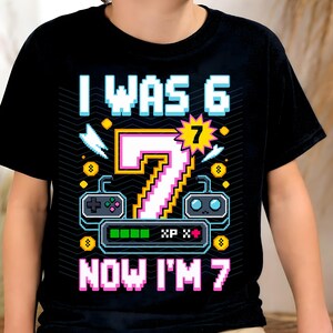 May include: Black t-shirt with a pixelated design celebrating a seventh birthday. The design includes the text "I WAS 6 NOW I'M 7" with a large number 7 in the center, along with game controller graphics and a progress bar.
