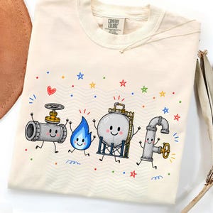 May include: A cream-colored t-shirt featuring a graphic of four animated characters representing the word HOPE: a grey pipe valve, a blue flame, a round storage tank, and a grey pipe with a wheel, surrounded by colorful stars and a red heart.