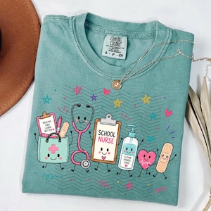 May include: A sage green t-shirt featuring a graphic print of a smiling medical bag, stethoscope, clipboard with the text 'SCHOOL NURSE', hand sanitizer bottle labeled 'CLEAN HANDS CARE', heart, and bandage, surrounded by colorful stars and hearts.