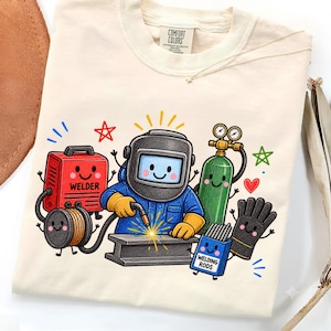 Op de afbeelding: Een crèmekleurig t-shirt met een cartoonafbeelding van een lasser in een blauw uniform en helm, een rood lasapparaat, een groene gasfles, een zwarte handschoen en een blauwe doos met het label WELDING RODS, allemaal met lachende gezichten.