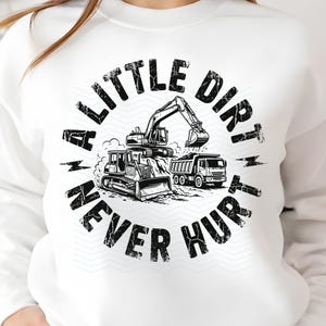 May include: White sweatshirt with a black and white graphic. The graphic features construction vehicles, including a bulldozer, excavator, and dump truck, with the text "A Little Dirt Never Hurt".