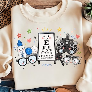 Könnte beinhalten: Ein cremefarbenes Sweatshirt mit einer bunten Grafik aus anthropomorphen Optometrie-Instrumenten, darunter eine Sehtafel mit den Buchstaben E, F, P, T, O, Z, L, P, E, D, P, E, C, F, D, E, D, F, C, Z, P, Augentropfen und Phoropteren.