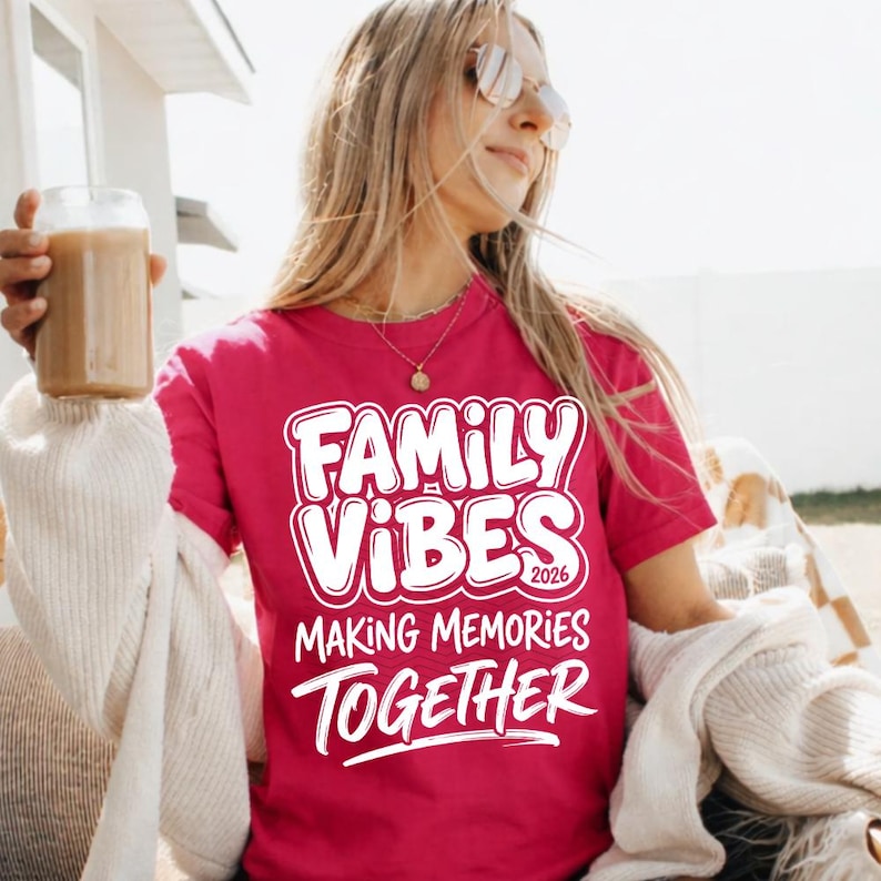May include: A magenta t-shirt with the words "FAMILY VIBES 2026 MAKING MEMORIES TOGETHER" in white, outlined text. The shirt is short-sleeved. A person is wearing the shirt and holding a glass of a brown beverage.