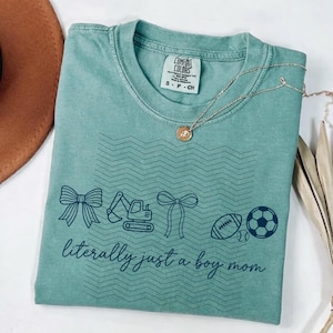 Puede incluir: Camiseta Comfort Colors verde azulado con cuello redondo, con un diseño de lazo, excavadora, lazo, fútbol americano, béisbol y balón de fútbol. El texto "literally just a boy mom" está impreso debajo. Un collar dorado está sobre la camiseta.