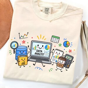 Puede incluir: Una camiseta de color crema con un estampado gráfico de un portátil sonriente con el texto DATA ANALYST en la pantalla. El diseño incluye ilustraciones de estilo dibujo animado de una base de datos, una carpeta, un cuaderno, una taza de café y una calculadora.