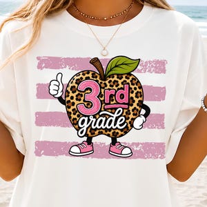 Puede incluir: Una camiseta blanca con un gráfico de una manzana con estampado de leopardo, una hoja verde, zapatillas rosas y guantes blancos. La manzana hace un gesto de pulgar hacia arriba y muestra el texto rosa y blanco 3rd grade sobre rayas rosas.