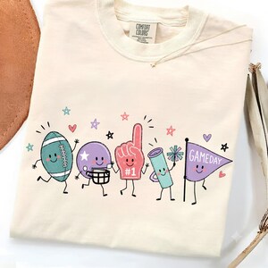 以下が含まれることがあります： クリーム色のTシャツで、フットボール、ヘルメットを被った紫色のボール、#1と書かれた赤いフォームフィンガー、メガホン、GAMEDAYと書かれた紫色のペナント旗など、漫画風のスポーツアイテムのグラフィックプリントが施されています。