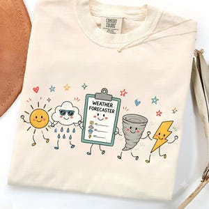 May include: A cream-colored t-shirt featuring a graphic of cartoon weather elements including a sun, a rain cloud with sunglasses, a clipboard labeled WEATHER FORECASTER, a tornado, and a lightning bolt, all surrounded by small colorful stars and hearts.