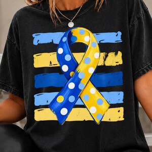 May include: A black t-shirt featuring a central awareness ribbon graphic. The ribbon is split vertically into blue and yellow sections, decorated with white and light blue polka dots. Behind the ribbon are horizontal brushstroke stripes.