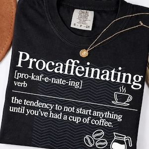 May include: Black t-shirt with the word "Procaffeinating" in white, along with a definition and coffee-related graphics. A gold necklace with a pendant rests on the shirt. The shirt has a Comfort Colors tag.