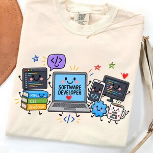 May include: A cream-colored t-shirt featuring a graphic of a smiling laptop with the text "SOFTWARE DEVELOPER" and a red heart. Surrounding elements include a stack of books labeled "HTML," "CSS," and "JavaScript," a coffee mug with "CODE COFFEE REPEAT" text, a smartphone, a monitor, and a blue gear icon.